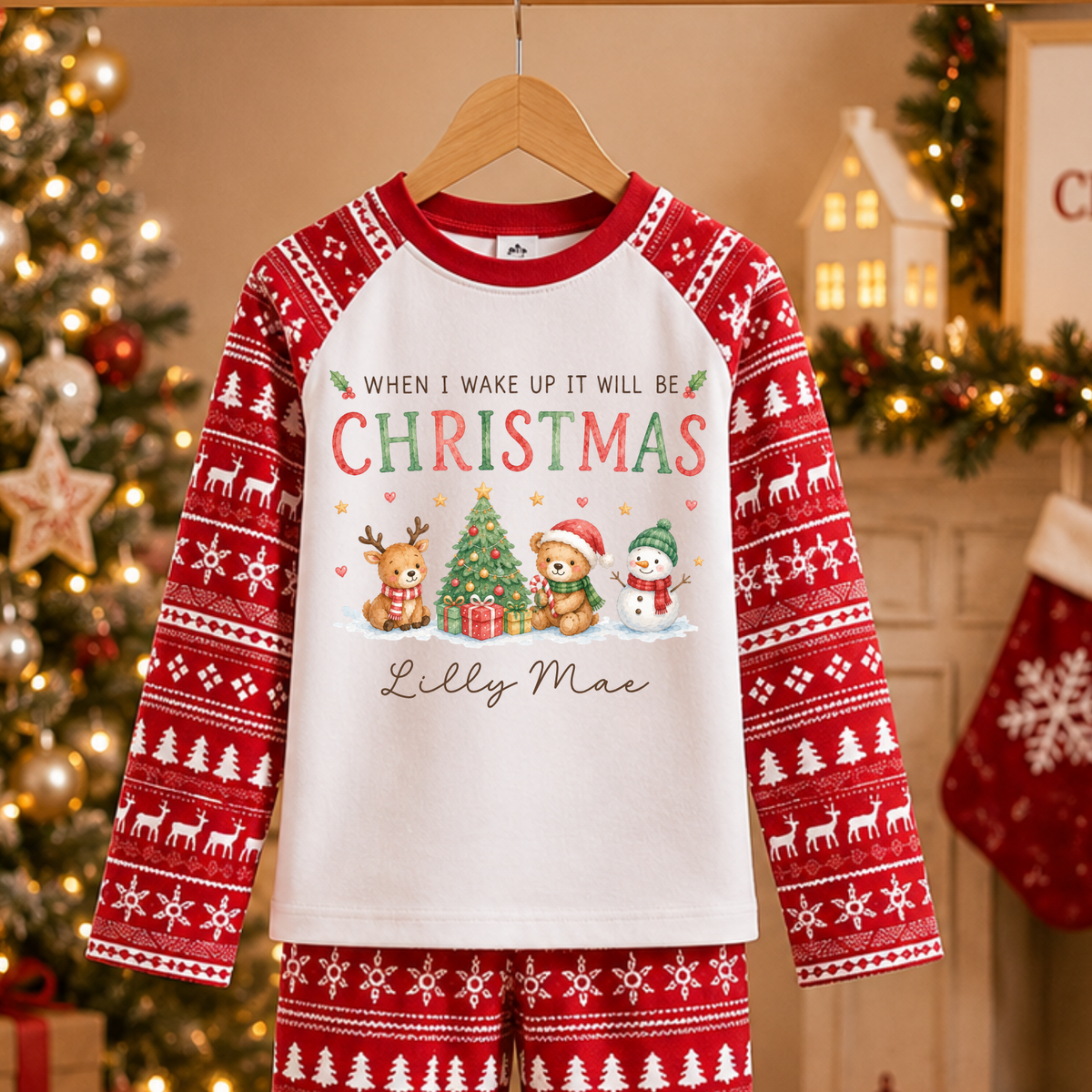 A two-piece Christmas pajama set hanging on a wooden hanger against a cozy holiday background. The set includes a long-sleeve top and matching pants in red and white, featuring a white front panel with the text 'WHEN I WAKE UP IT WILL BE CHRISTMAS' above a festive illustration of a tree, gifts, Santa, and a snowman, with 'Lilly Mae' printed below.