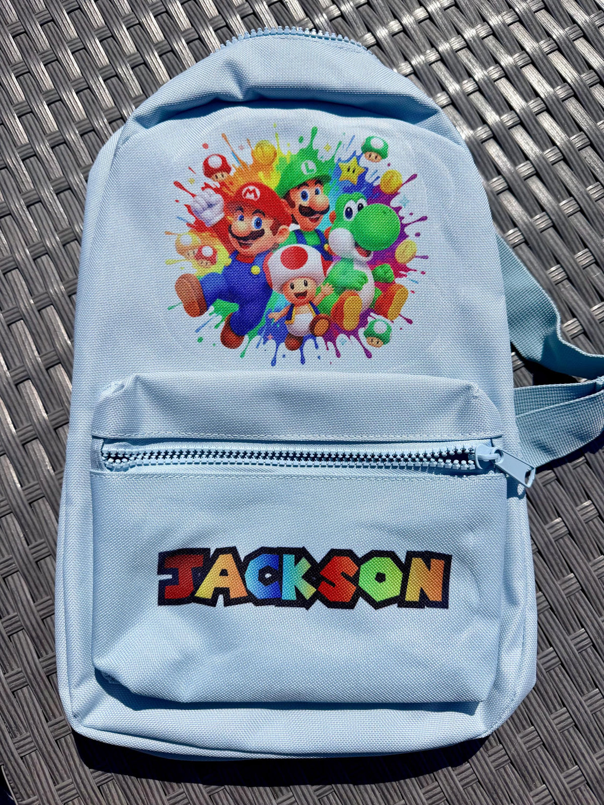 A light blue backpack featuring a colorful Mario-themed character graphic on the upper front and the name 'JACKSON' in a multicolor pixel-style font on the lower front pocket. The backpack is photographed from above on a dark gray woven patio surface.