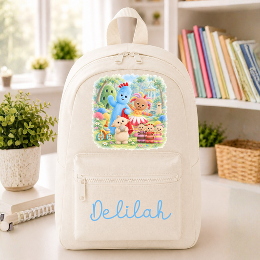 A cream-colored canvas backpack featuring a colorful cartoon illustration of characters from 'The Big Winged Sun' on the front, with the name 'Delilah' personalized in light blue script on the front pocket. The backpack is displayed on a tabletop in a bright, cozy room with a bookshelf and potted plants in the background.