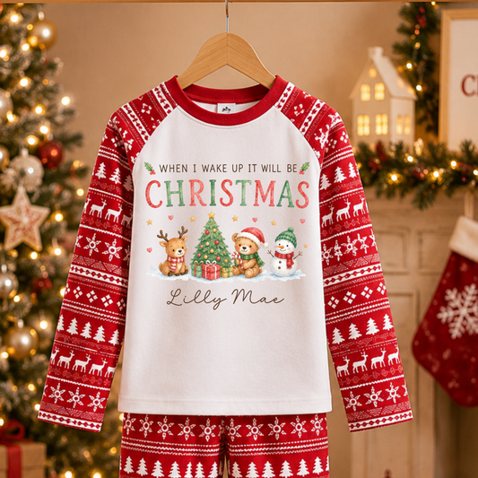 A two-piece Christmas pajama set hanging on a wooden hanger against a cozy holiday background. The set includes a long-sleeve top and matching pants in red and white, featuring a white front panel with the text 'WHEN I WAKE UP IT WILL BE CHRISTMAS' above a festive illustration of a tree, gifts, Santa, and a snowman, with 'Lilly Mae' printed below.