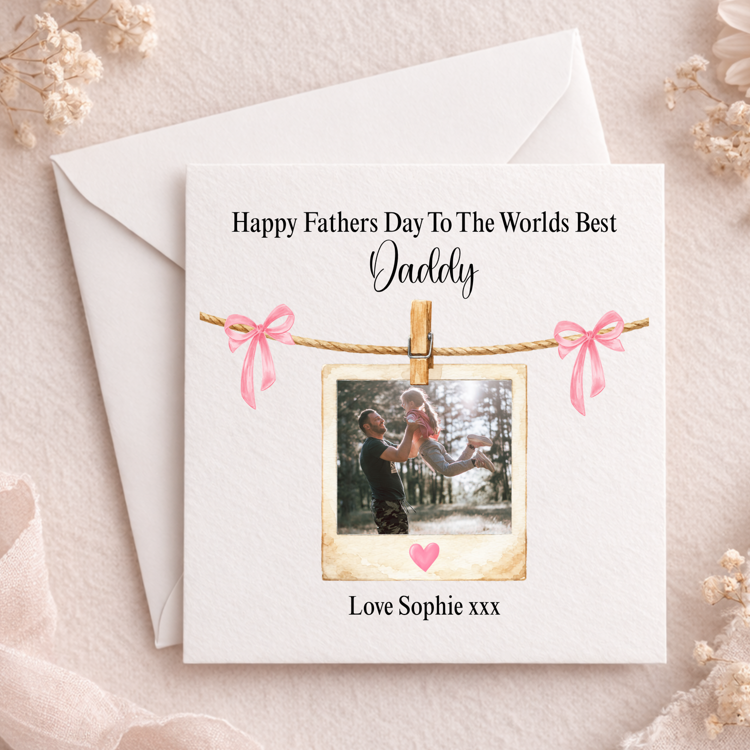 Father's Day card with photo of a man and child, pink ribbons, and envelope on a light background.