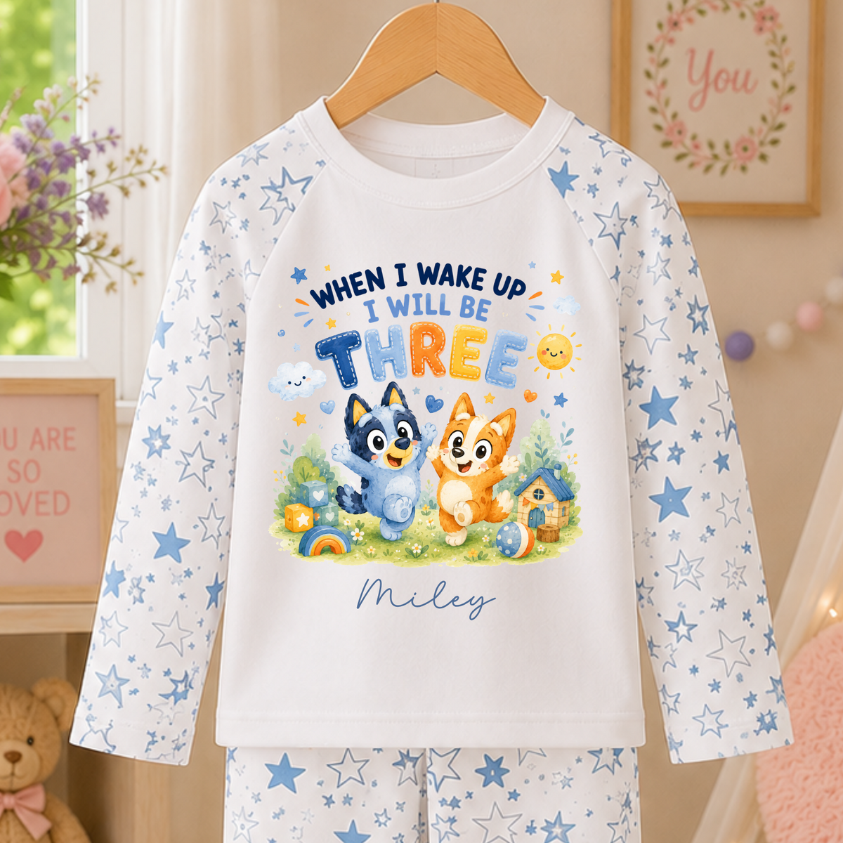 A children's long-sleeve pajama set hanging on a wooden hanger, featuring a white top with blue star prints and a colorful cartoon graphic that reads 'WHEN I WAKE UP I WILL BE THREE' with the name 'Miley' below, paired with matching white pants with blue star prints. The background is a cozy, warm room setting with a teddy bear, framed art, and soft lighting.