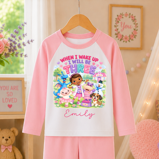 A children's two-piece pajama set hanging on a wooden hanger, featuring a long-sleeve top and matching pants. The pajama set has a white body with pink sleeves and a colorful cartoon graphic on the chest that reads 'WHEN I WAKE UP I WILL BE THREE' above the name 'Emily'.