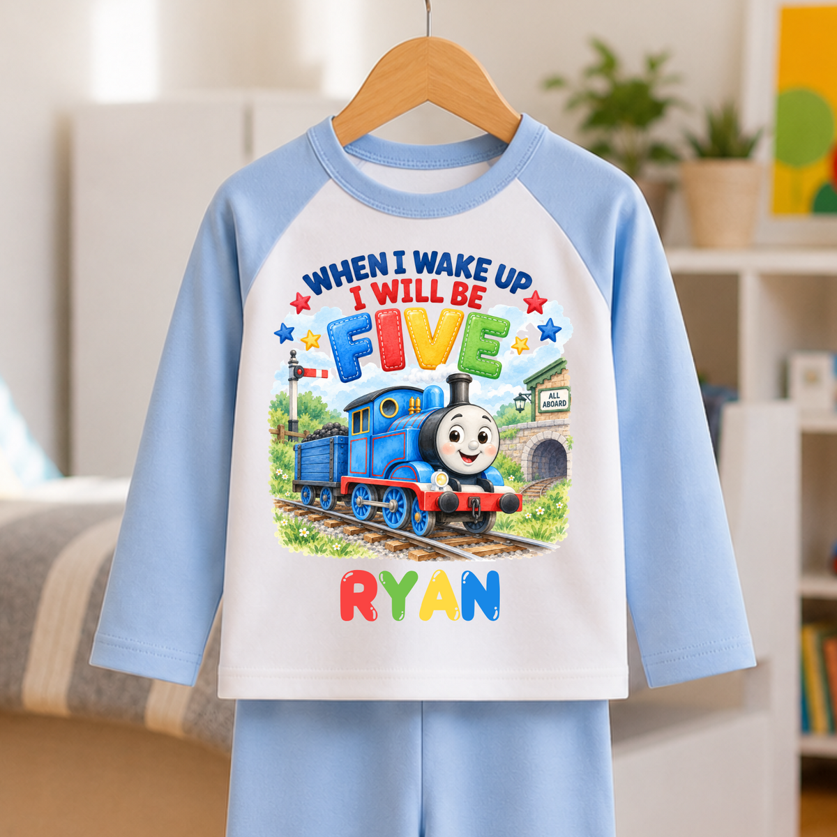 A children's two-piece pajama set hanging on a wooden hanger, featuring a light blue and white raglan long-sleeve top and matching light blue pants. The front of the top has a colorful graphic of Thomas the Tank Engine with the text 'WHEN I WAKE UP I WILL BE FIVE' and the name 'RYAN' in colorful letters.