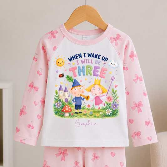A children's two-piece pajama set hanging on a wooden hanger, featuring a white long-sleeve top with pink bow and heart prints on the sleeves and a colorful fantasy graphic on the front, paired with matching pink pants with bow and heart patterns.