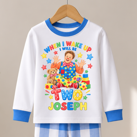 A children's long-sleeve pajama top hanging on a wooden hanger, paired with blue-and-white check pajama pants. The top is white with blue trim and a colorful cartoon clown graphic on the front that reads 'WHEN I WAKE UP I WILL BE TWO JOSEPH.