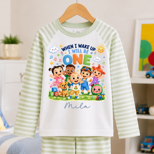 A children's two-piece pajama set hanging on a wooden hanger, featuring a white and light green striped long-sleeve top and matching striped pants. The top has a colorful graphic print with cartoon children and the text 'WHEN I WAKE UP I WILL BE ONE' and 'Mila'.
