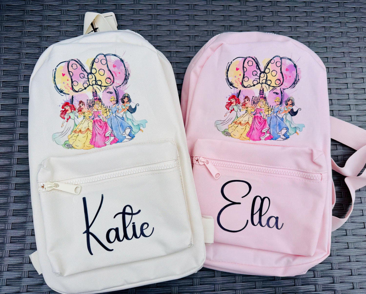 Two personalized canvas backpacks side by side on a dark woven mat. The left backpack is off-white/cream and personalized with the name 'Katie' in black script, featuring a colorful graphic of Disney princesses under a Minnie Mouse-style bow.