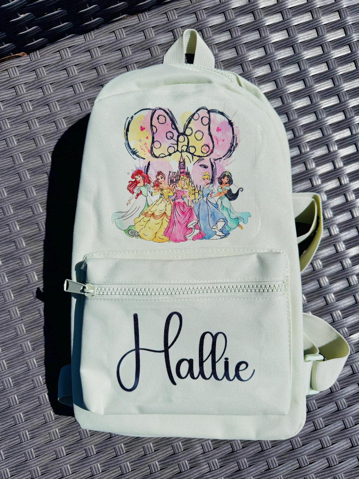 A personalized off-white canvas backpack with a colorful Disney princess graphic and the name 'Hallie' printed in black script on the front pocket, resting on a dark gray woven patio surface.