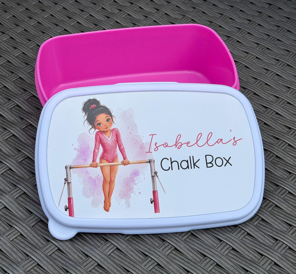 A custom pink and white plastic chalk box with a gymnast illustration and the text 'Isobella's Chalk Box' on the lid, shown open with its pink interior on a dark woven mat.