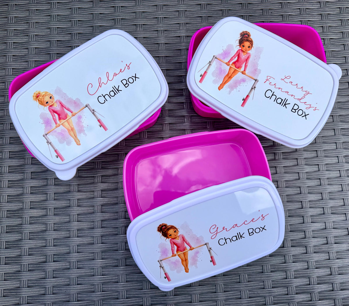 Three customisable pink plastic lunch boxes with white lids are arranged on a grey woven mat. Each lid features a watercolor illustration of a gymnast and the text 'Chloe's Chalk Box', 'Lenny's Funniest Chalk Box', and 'Grace's Chalk Box' respectively.