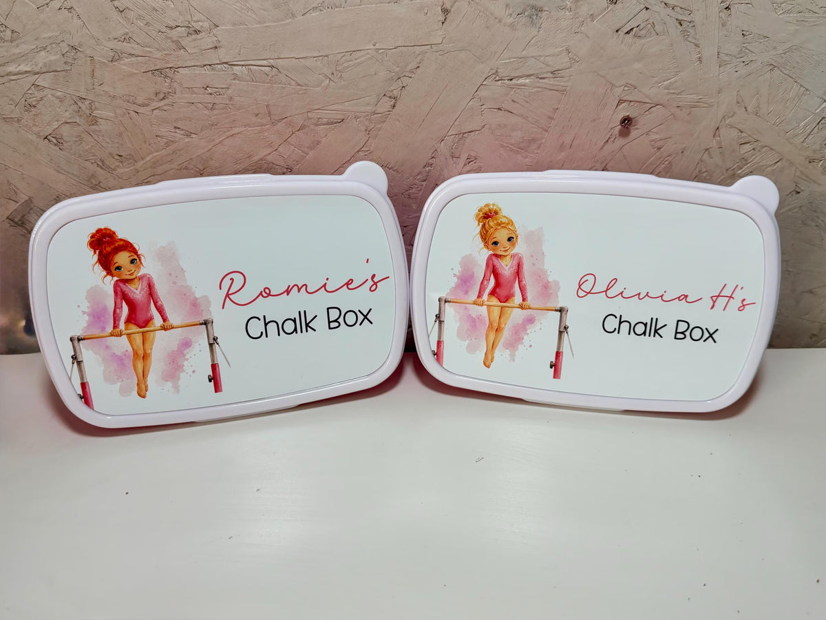 Two personalized chalk boxes with white lids featuring cartoon gymnast illustrations and the text 'Romie's Chalk Box' and 'Olivia's Chalk Box', displayed on a white surface against a textured brown background.
