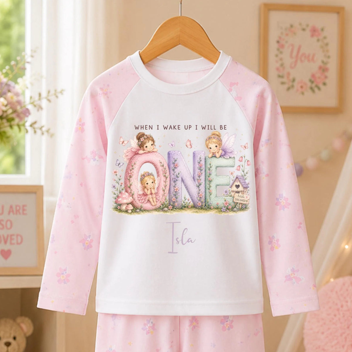 A children's two-piece pajama set hanging on a wooden hanger, featuring a long-sleeve top and matching pants in soft pink and white. The top has a large front graphic with the word 'ONE' in pastel letters and cute cartoon characters, with the text 'WHEN I WAKE UP I WILL BE' above it.