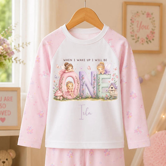 A children's two-piece pajama set hanging on a wooden hanger, featuring a long-sleeve top and matching pants in soft pink and white. The top has a large front graphic with the word 'ONE' in pastel letters and cute cartoon characters, with the text 'WHEN I WAKE UP I WILL BE' above it.