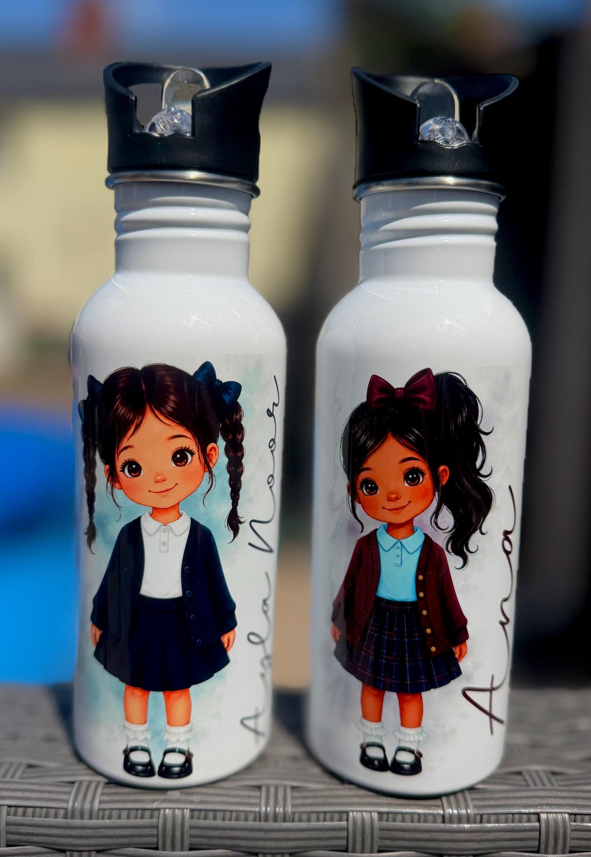 Two white insulated water bottles with black flip-top lids, each printed with a cartoon girl in school uniform. The bottles are standing side by side on a woven gray surface with a blurred outdoor background.