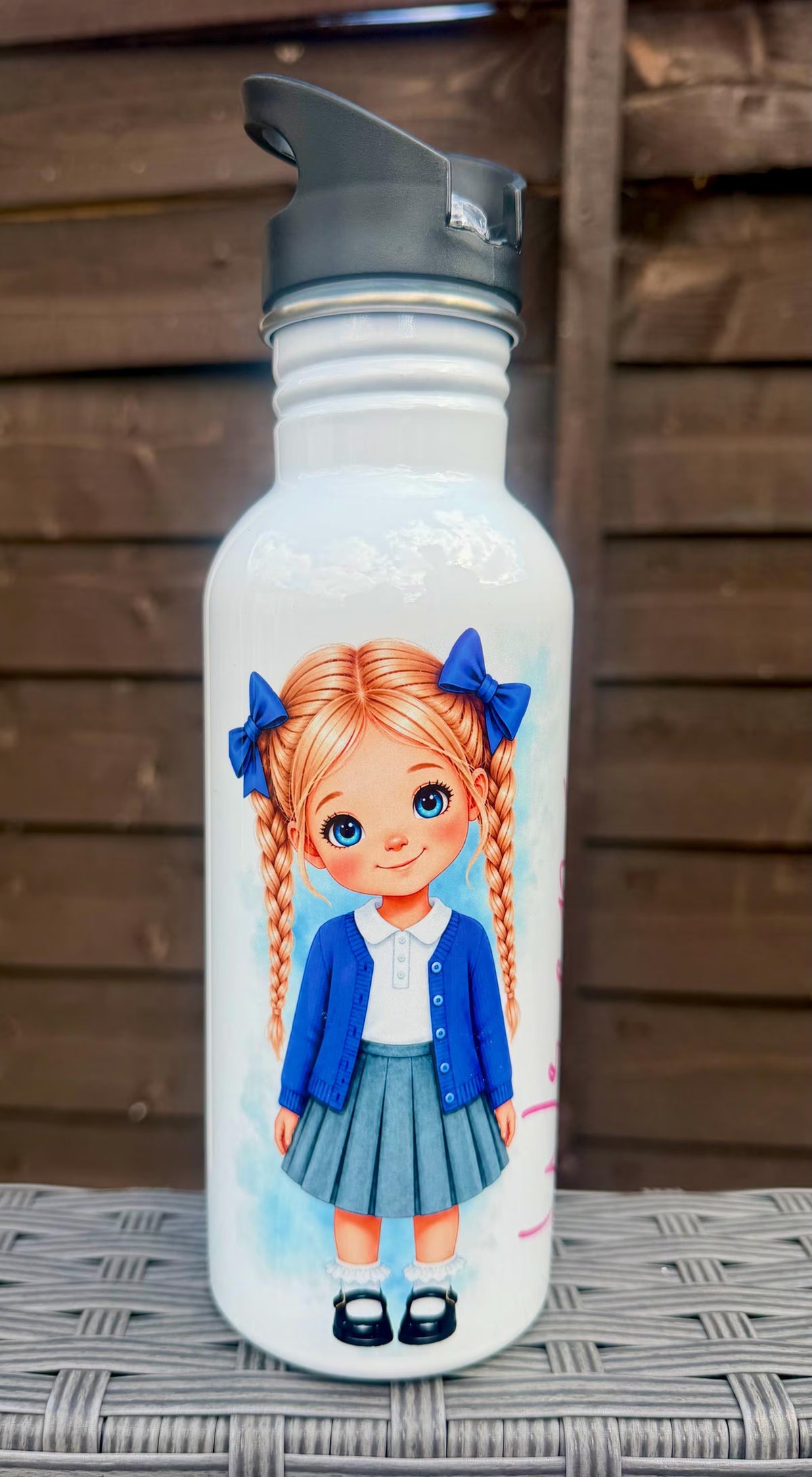 A white insulated water bottle with a black flip-top lid, featuring a printed illustration of a girl with orange pigtails and blue bows, standing on a gray woven surface against a dark wooden background.