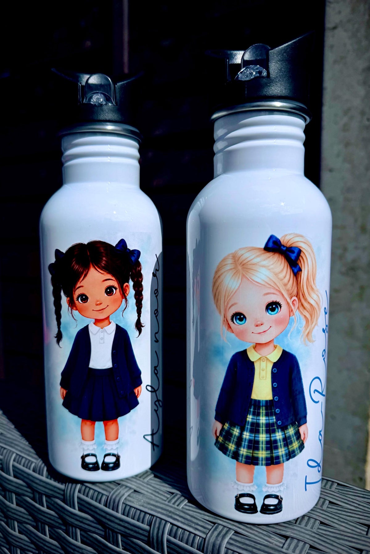 Two white insulated water bottles with black flip-top lids, each printed with a cartoon girl in school uniform. One bottle shows a dark-haired girl with braids, and the other shows a blonde girl with a ponytail.