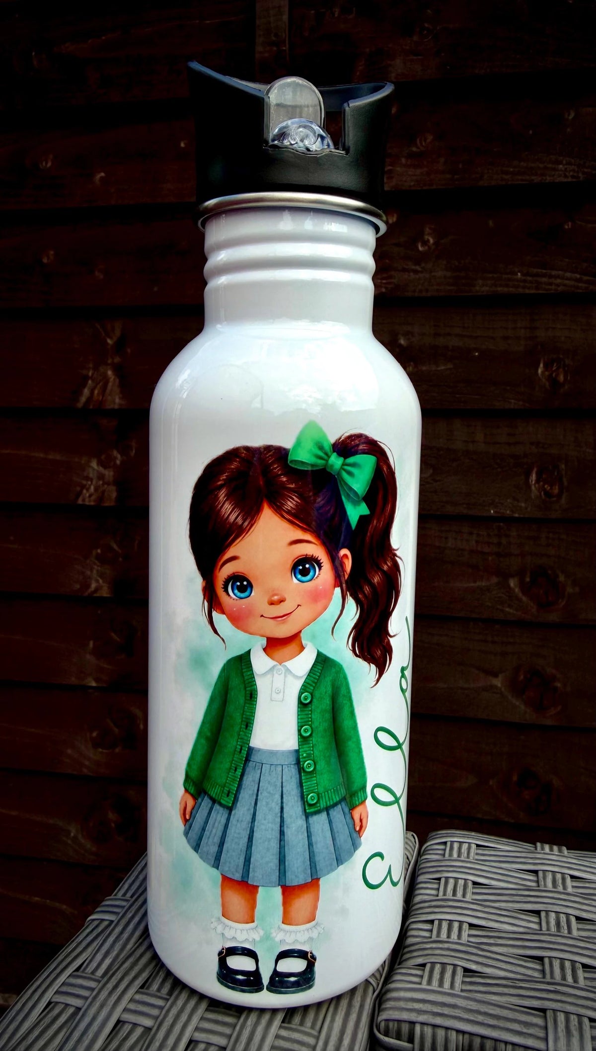 A white insulated water bottle with a black flip-top lid, featuring a printed illustration of a girl with brown hair in a green bow, wearing a green cardigan and blue pleated skirt. The bottle is photographed on a woven surface against a dark wooden plank background.